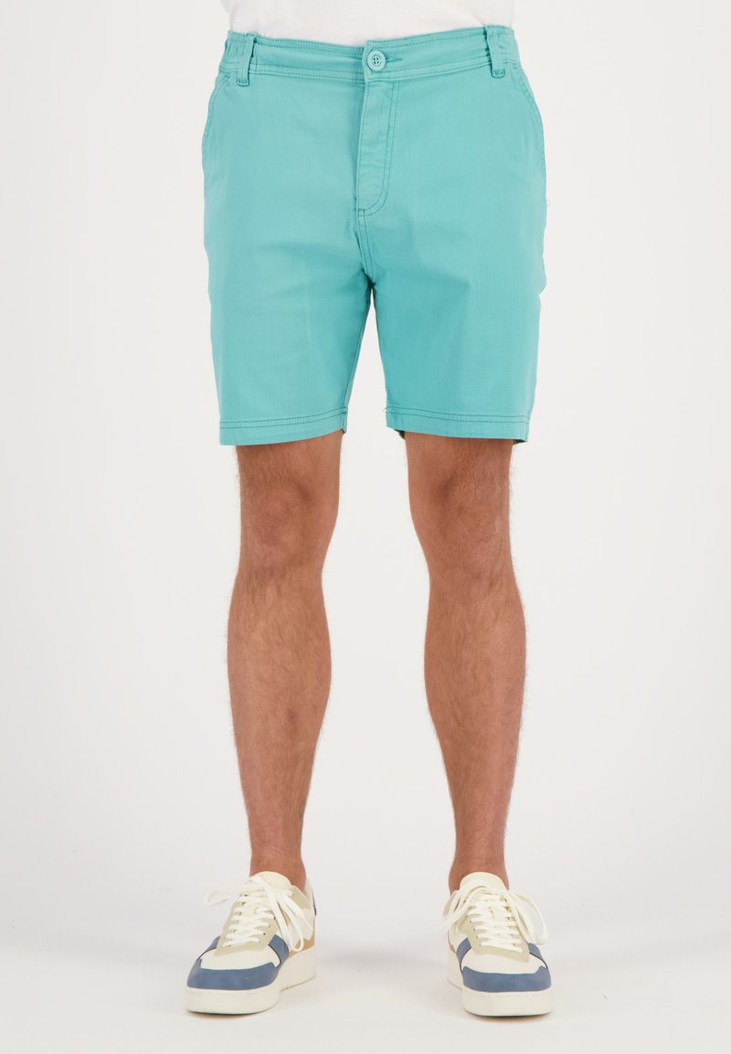 Pantalones cortos turquesa de algodón, con un diseño de frente plano, bolsillos estándar y cierre de botón. Combinados con zapatillas de varios colores.