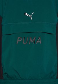 Chaqueta ligera en tono azul verdoso con un bolsillo frontal, detalles en negro, un logo de PUMA en impresión texturizada y un detalle de logo a rayas en contraste.