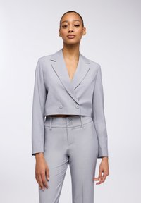 Blazer grigio accorciato con doppi bottoni, colletto a notched e maniche lunghe, abbinato a pantaloni su misura coordinati con vita alta e doppi bottoni.