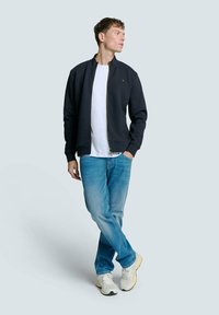 Marine-blue zip-up jack met een gestructureerd patroon, wit T-shirt, lichtblauwe spijkerbroek en crèmekleurige sneakers. De jack heeft een opstaand kraag en logodetail.