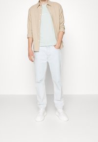 Beige overhemd met knopen over een lichtblauw T-shirt met witte tekst, gecombineerd met lichtblauwe jeans en witte sneakers. Eenvoudig ontwerp, losse pasvorm.