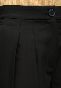 Μαύρο παντελόνι ψηλόμεσο με πιέτες και ένα μαύρο κουμπί στη μέση, μπροστά σε μπεζ υφή φόντο.