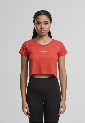 Piros, rövidre szabott pamut póló, kerek nyakkivágással és rövid ujjakkal, az elülső részen fehér logóval. Fekete leggings-szel párosítva.