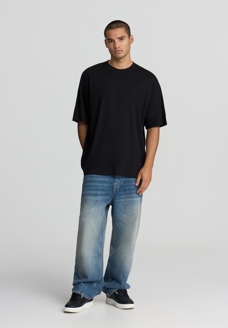 T-shirt nera in cotone a maniche corte, abbinata a jeans a gamba larga blu chiaro sbiaditi e sneakers nere, in piedi contro uno sfondo neutro.