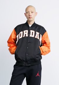 Bald person wearing a black and orange Jordan varsity jacket and black pants with a red Jumpman logo, hands in pockets, neutral expression.
