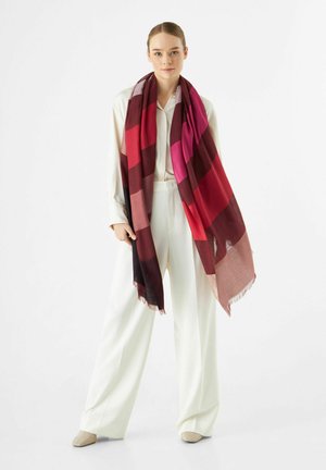 Grand foulard rectangulaire aux nuances dégradées de rouge, rose et bordeaux. Fabriqué en tissu léger avec des bords frangés, drapé sur une tenue blanche.