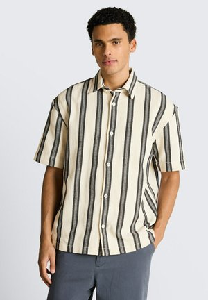 Mann trägt cremefarbenes Kurzarmhemd mit vertikalen schwarzen Streifen und graue Hose, steht vor schlichtem hellgrauem Hintergrund.