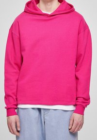 Felpa rosa brillante realizzata in tessuto morbido, con un taglio rilassato, grande cappuccio e polsini a costine. Indossata sopra una t-shirt bianca, abbinata a jeans chiari.