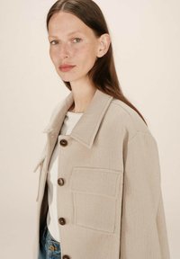 Kvinde med langt brunt hår iført en beige knapjakke over en hvid skjorte og blå jeans, der vender let til venstre foran en ensfarvet baggrund.