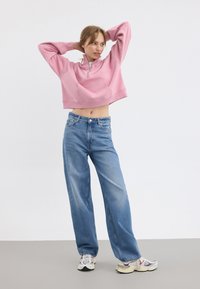 Korte roze sweatshirt met een voorzak en halve rits, gecombineerd met lichtblauwe wijde jeans en kleurrijke sneakers.
