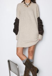 Robe beige à manches longues avec un ourlet côtelé, ornée de manches imitant le cuir marron foncé. Portée avec des bottes marron jusqu'au genou et des chaussettes grises.