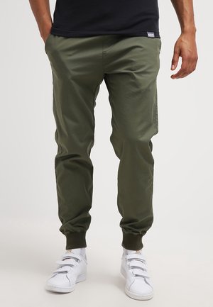 Mann trägt olivgrüne Jogginghosen mit elastischen Bündchen, weiße Turnschuhe und ein schwarzes Hemd, steht entspannt mit den Händen.