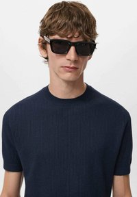Occhiali da sole neri con montatura rettangolare, indossati da una persona in un maglione corto a maniche corte blu navy a coste, su uno sfondo neutro.