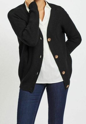 Cardigan nero lavorato a maglia con scollo a V, cinque grandi bottoni di legno e trama a coste, abbinato a una camicetta bianca e jeans blu.