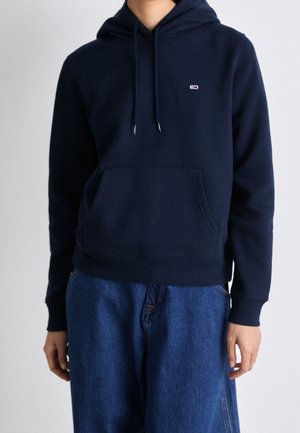 Persoon draagt een marineblauwe hoodie met trekkoorden en een voorzak, gecombineerd met loszittende blauwe spijkerbroeken.