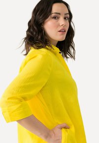 Camicia gialla realizzata in tessuto leggero con colletto alla mandarina e maniche a tre quarti. Presenta una vestibilità comoda e una tasca laterale.