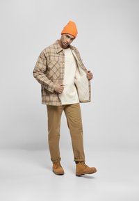 Kurtka w kratę w odcieniach brązu, beżowa koszula pod spodem, khaki spodnie, pomarańczowa czapka beanie i brązowe buty. Teksturowany i warstwowy design z widocznym przeszyciem.
