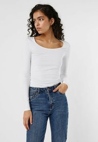 Weißes Langarmshirt mit runder Ausschnitt, figurbetontem Design, weicher Textur, kombiniert mit hochtaillierten blauen Jeans.