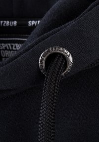 Detail van een zwarte hoodie die een ronde metalen oogje toont met de tekst "SPITZBUB," met een textuurkoord in zwart en zachte stof eromheen.