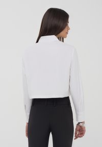 Blusa bianca cropped con collare, maniche lunghe e polsini con bottone, abbinata a pantaloni neri aderenti. Tessuto liscio, design minimalista.