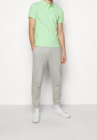 Muž ve světle zeleném polo tričku, šedých teplácích, bílých ponožkách a bílých teniskách stojí uprostřed prosté bílé zdi.