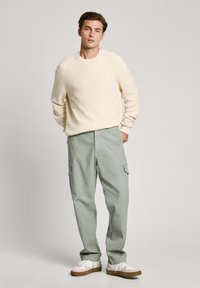 Krem pulover z rebrastim vzorcem in svetlo zelenimi cargo hlačami. Cargo hlače imajo stranske žepe. Bele športne copate z rjavimi podplati. Nevtralna kulisa.