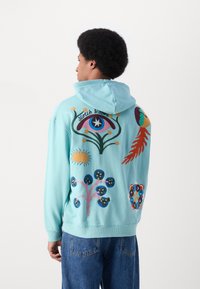 Felpa azzurra chiara con disegni ricamati colorati, tra cui un occhio, fiori e forme astratte sul retro. Tessuto morbido, vestibilità comoda.