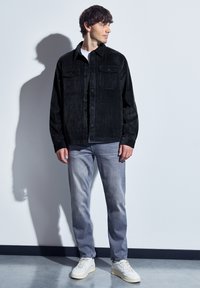 Černá kordurová košile s knoflíkovým zapínáním, dvěma náprsními kapsami, ve spojení s světlými šedými džíny a bílými teniskami. Vnitřní prostředí.