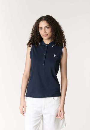 Donna con capelli ricci che indossa una polo senza maniche blu navy con rifiniture bianche e pantaloni bianchi, in piedi contro uno sfondo chiaro e semplice.