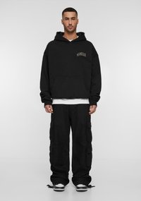 Felpa nera con cappuccio, tasca frontale e logo, abbinata a pantaloni cargo neri con tasche laterali. Le sneakers completano il look.