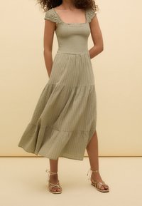 Robe midi vert clair avec un décolleté carré, des manches volantes, un tissu texturé et une jupe à volants. Associée à des sandales dorées à lanières.