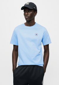 Ανοιχτό μπλε βαμβακερό μπλουζάκι με στρογγυλή λαιμόκοψη και κοντά μανίκια, με κυκλικό ένθετο στην α ριστερή πλευρά του στήθους. Μαύρο αθλητικό παντελόνι.