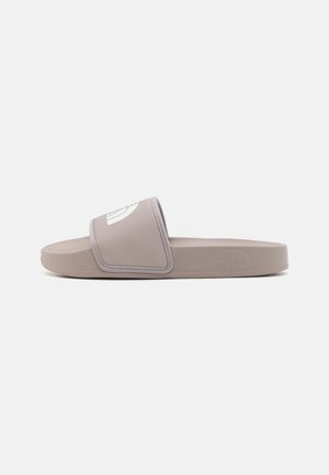 Sandalia tipo slide en suave goma gris. Cuenta con una tira ancha con un logo blanco, superficie lisa, suela plana y plantilla contorneada para mayor comodidad.