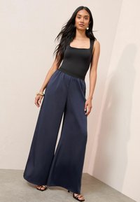 Frau mit langen dunklen Haaren, die ein schwarzes ärmelloses Oberteil, weite marineblaue Hosen und schwarze Sandalen mit Absatz trägt, steht vor einer beigen Wand.