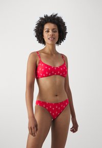 Rdeč bikini set, ki vključuje top z tankimi naramnicami in nizkimi boksericami. Obe delu imata bel vzorec sidra na enotni rdeči podlagi.