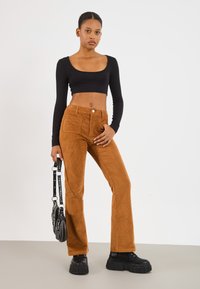 Barna corduroy, harangszárú nadrág elülső zsebekkel, egy fekete, hosszú ujjú cropped toppal párosítva. A modell egy fekete kézitáskát tart, amelynek pántja van.