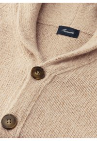 Beige wollen cardigan met een sjawlboord, twee bruine knopen en een geweven structuur. Label genaaid aan de binnenkant bij de kraag.