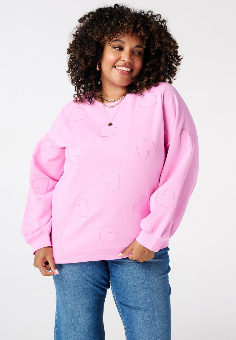 Glunderende vrouw met krullend haar, gekleed in een roze sweatshirt met gestikte harten en blauwe spijkerbroek, poserend tegen een effen witte achtergrond.