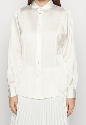 Woman wearing a long-sleeve white satin button-up shirt with a collar and a white pleated skirt, standing against a plain background.