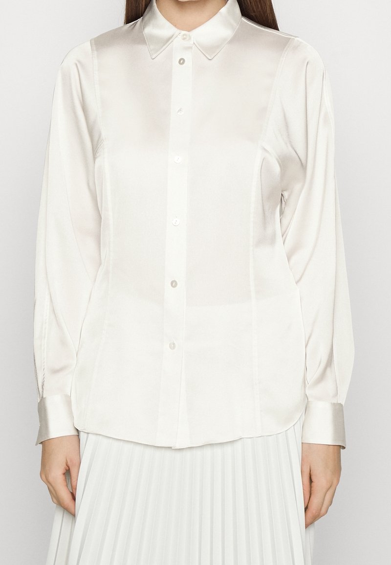 Femme portant une chemise blanche en satin à manches longues avec col et une jupe plissée blanche, debout devant un fond uni.