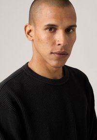 Black textured sweater with a round neckline, featuring a ribbed knit design. Model has short hair and is facing the camera.