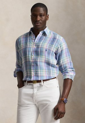 Vyras, dėvintis pastelinių spalvų languotą marškinių viršų su įgulėtomis rankovėmis, baltas kelnes, rudą diržą ir rudą odinį laikrodį ant riešo.