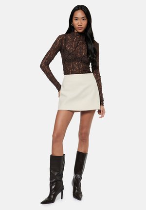 Kvinde iført en brun blondebluse med lange ærmer, en cremefarvet mininederdel og sorte knæhøje støvler med høje hæle, stående med den ene hånd på hoften.