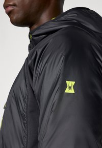 Melna izolēta jaka ar kapuci, ar gludu tekstūru, dzeltenu zirneklīša logo uz piedurknes un kontrastējošu iekšējo krāsu.