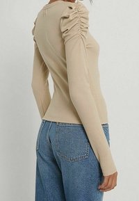 Langärmliges, geripptes Oberteil in Beige mit gerafften Schulterakzenten; taillierte Passform. Kombiniert mit hochgeschnittenen blauen Denim-Jeans.