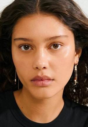 Orecchini in tono oro con catene pendenti, caratterizzati da una combinazione di perline rotonde bianche, arancioni e viola scuro, indossati su una camicia nera.