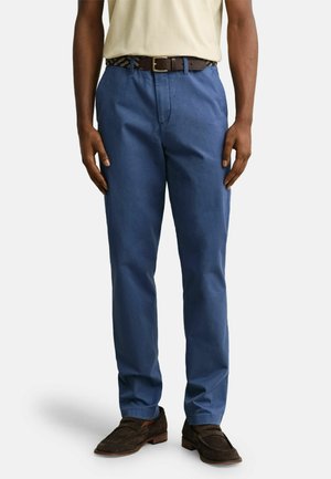 Man draagt blauwe rechte broek, bruine riem, beige overhemd in de broek gestopt en bruine suède loafers, staand op een witte achtergrond.