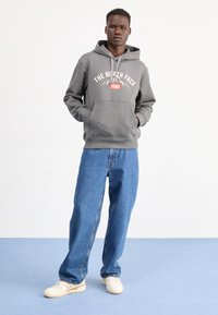 Szara bluza z kapturem z frontową grafiką, sznurkiem w kapturze, bocznymi kieszeniami, w parze z jasnoniebieskimi dżinsami i białymi sneakersami na niebieskiej podłodze.