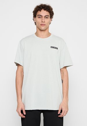 Jeune homme aux cheveux bouclés portant un t-shirt gris clair uni avec un petit logo noir sur le haut gauche de la poitrine, debout devant un fond blanc.