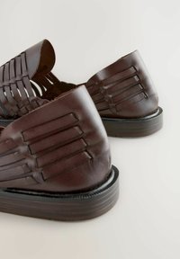 Sandalias de cuero marrón con un diseño superior trenzado, suela plana y detalles horizontales a lo largo del talón. Textura suave con un acabado pulido.